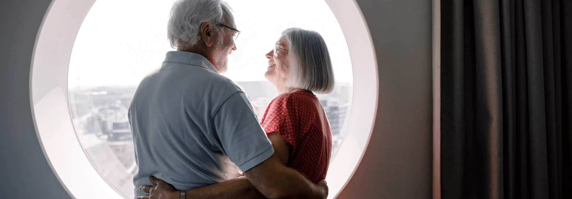 An elderly couple stand in front of a circular window, smiling at each other.