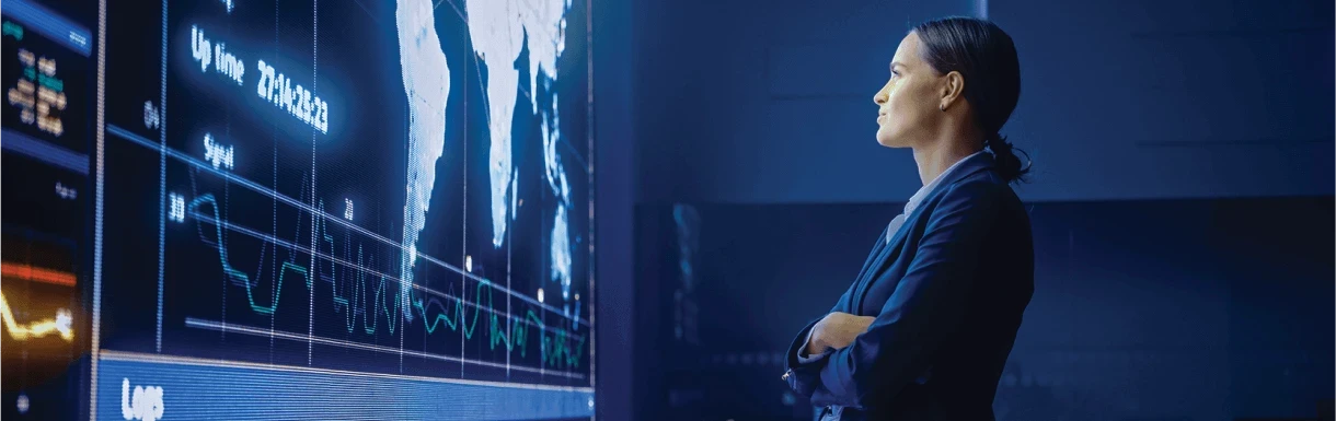 A woman wearing a dark blue suit stands in front of a wall-size display screen, viewing performance log data on a world map.