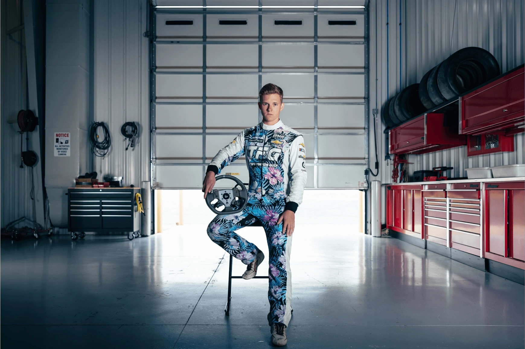 Professional stock car driver Jack Wood wears his racing uniform and sits on a stool in a garage holding a Logitech G PRO Wheel.