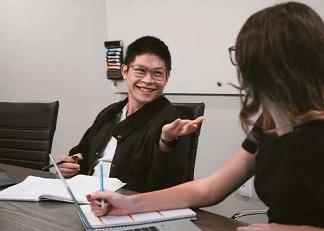 Senior Media Associate Quan Pham smiling and gesturing at team member Digital Marketing Associate Katie Anderson sitting beside him at a conference table with open notebooks and pens poised for note-taking in Adcetera’s Chicago office, a Digital Advertising Agency.