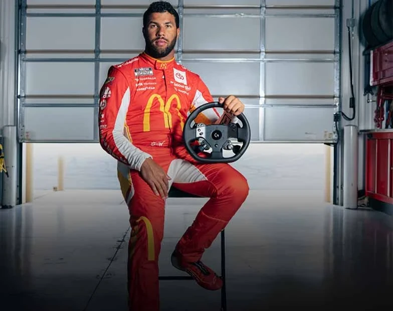 Professional stock car driver William Darrell “Bubba” Wallace wears his red racing uniform and sits on a stool in a garage holding a Logitech G PRO Wheel.