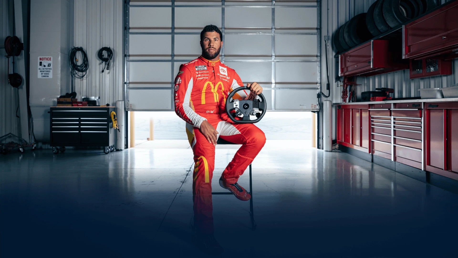 Professional stock car driver William Darrell “Bubba” Wallace wears his red racing uniform and sits on a stool in a garage holding a Logitech G PRO Wheel.