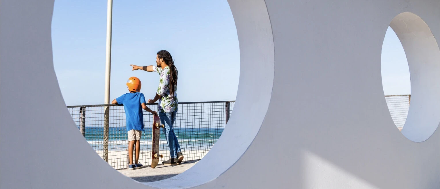 A man and a boy are framed by a circular opening in a wall, standing by a railing looking out toward the ocean.