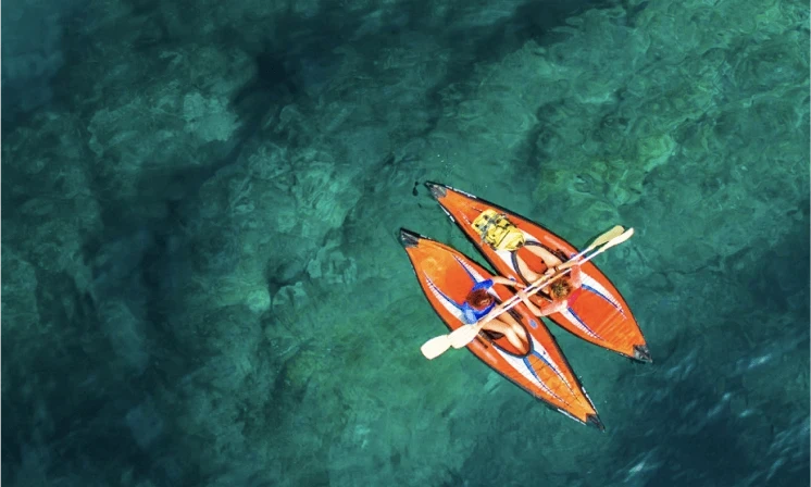 An overhead view of two orange kayaks floating side by side on turquoise water; the kayakers holding hands.